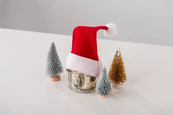 Маленькі сосни біля скляної банки з доларовими банкнотами з капелюхом Санта на білому — стокове фото