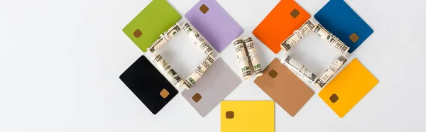 Panoramic shot of multicolored credit card templates near cash rolls on white — Stock Photo