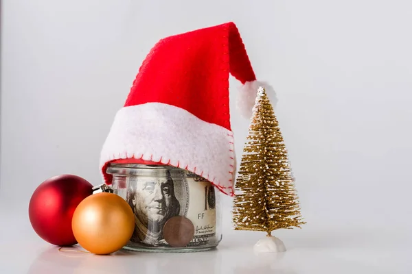 Небольшая елка возле рождественских балов и банка с долларовыми купюрами на белом — стоковое фото