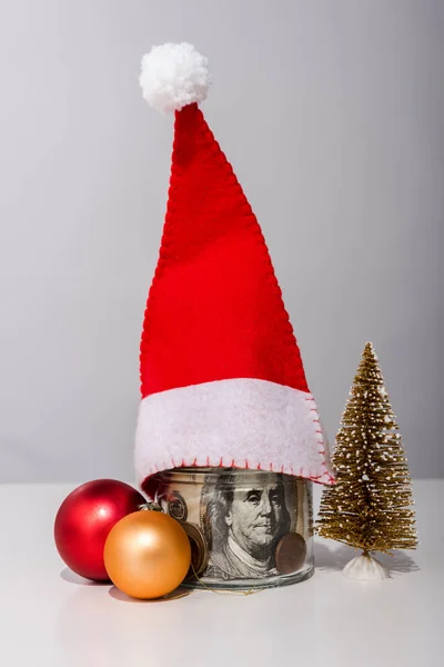 Boules et bocal de Noël avec billets en dollars et chapeau de Père Noël sur blanc — Photo de stock