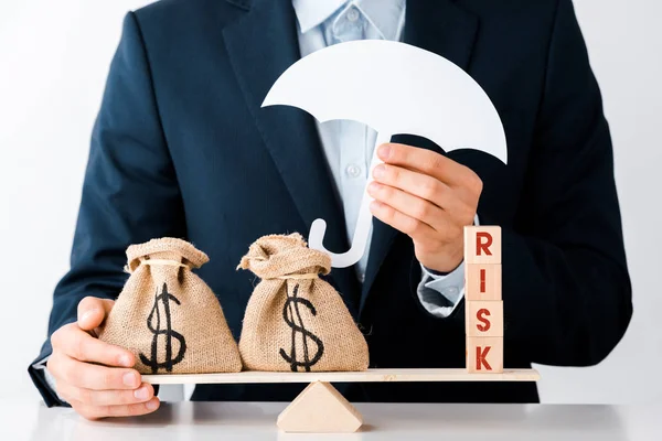 Vista cortada de empresário segurando guarda-chuva de papelão perto de balanças com sacos de dinheiro e blocos de madeira com letras de risco no branco — Fotografia de Stock