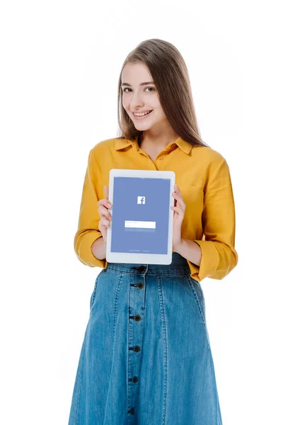 KYIV, UCRANIA - 12 de agosto de 2019: niña sonriente en falda de mezclilla sosteniendo tableta digital con aplicación de Facebook aislada en blanco - foto de stock