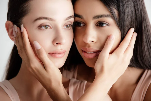 Beautiful young multicultural girls in beige outfit tenderly touching each other faces isolated on white — Stock Photo