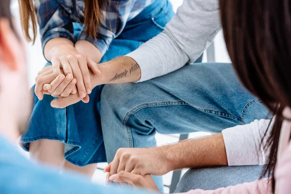 Vista recortada del grupo de apoyo cogido de la mano mientras está sentado en sillas aisladas en blanco - foto de stock