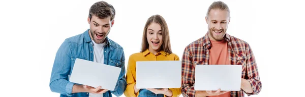 Felices jóvenes amigos sosteniendo computadoras portátiles con bocas abiertas aisladas en blanco, tiro panorámico - foto de stock