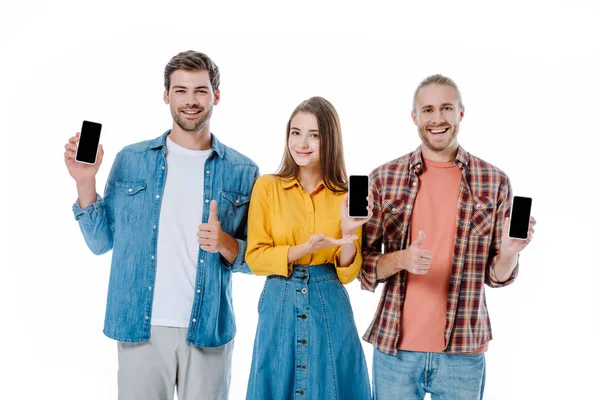 Tres jóvenes amigos sosteniendo teléfonos inteligentes con pantallas en blanco y mostrando los pulgares hacia arriba aislados en blanco - foto de stock