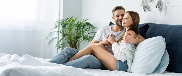 Panoramic shot of handsome man holding gift box and hugging smiling woman — Stock Photo