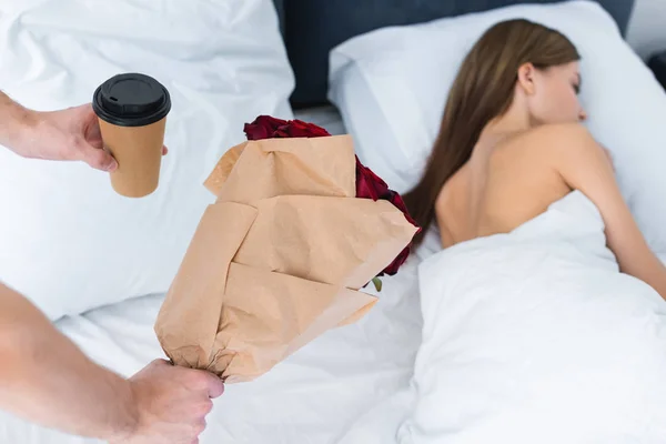 Vista cortada do homem segurando copo de papel, buquê e mulher dormindo na cama — Fotografia de Stock