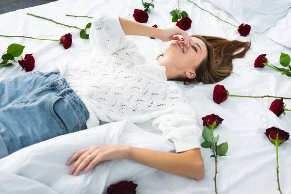 Sonriente mujer oscureciendo la cara y acostada en la cama con rosas - foto de stock