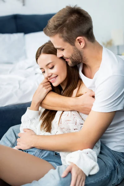 Hombre guapo abrazando mujer atractiva y sonriente en el apartamento - foto de stock