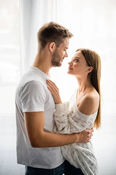 Vista lateral del hombre guapo abrazando a mujer atractiva y sonriente en el apartamento - foto de stock