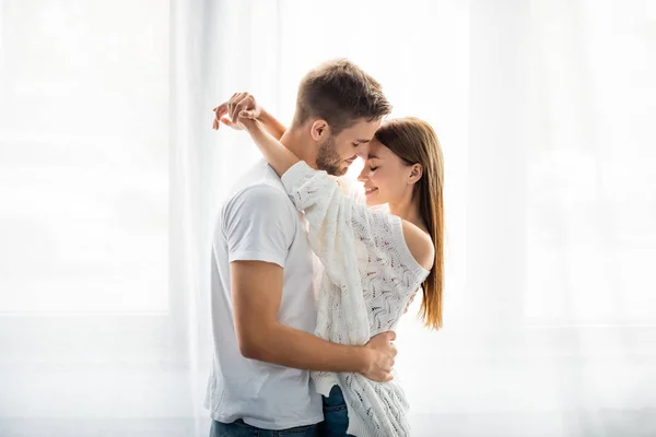 Vista laterale di bell'uomo che abbraccia donna attraente e sorridente in appartamento — Foto stock