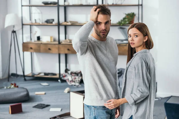 Attraktive Frau und schockierter Mann blicken in ausgeraubte Wohnung in die Kamera — Stockfoto