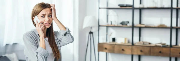 Panoramic shot of attractive and sad woman talking on smartphone in robbed apartment — Stock Photo