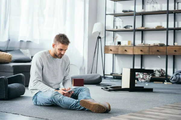 Schöner Mann sitzt auf dem Boden und benutzt Smartphone in beraubter Wohnung — Stockfoto