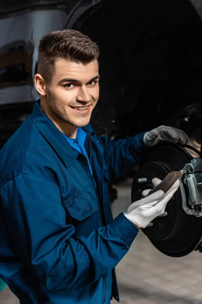 Junger Mechaniker schaut in Kamera, während er Bremsbelag hält — Stockfoto