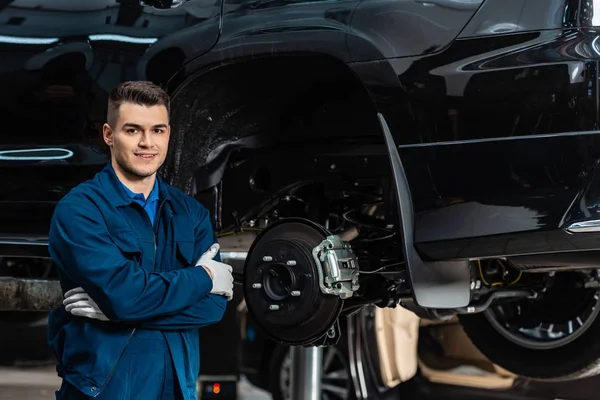 Lächelnder Mechaniker blickt in die Kamera, während er mit verschränkten Armen in der Nähe montierter Scheibenbremsen steht — Stockfoto