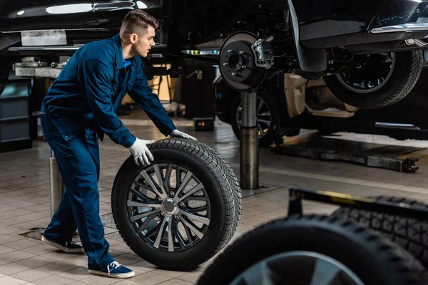 Joven mecánico sosteniendo rueda de coche cerca de coche levantado en taller - foto de stock