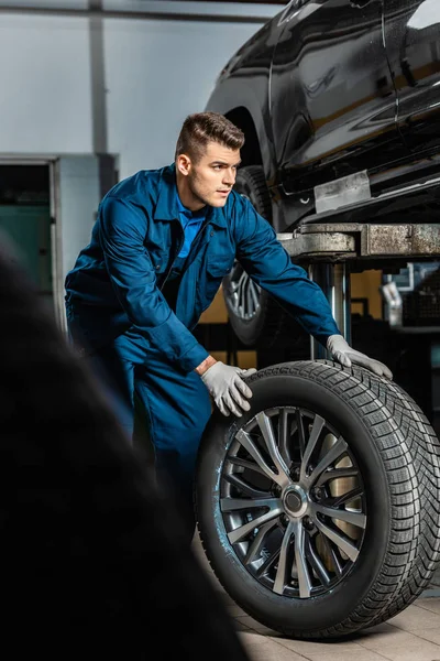 Selective focus of young mechanic with car wheel near raised car in workshop — Stock Photo