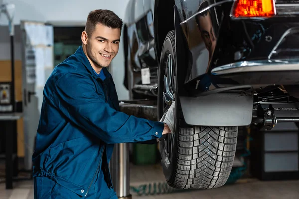 Giovane meccanico che installa ruota su auto sollevata in officina e sorride alla macchina fotografica — Foto stock