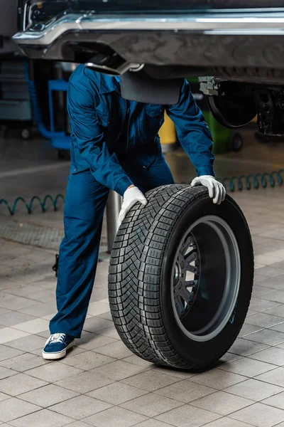 Обрезанный вид механика с колесом рядом с поднятым автомобилем в мастерской — стоковое фото