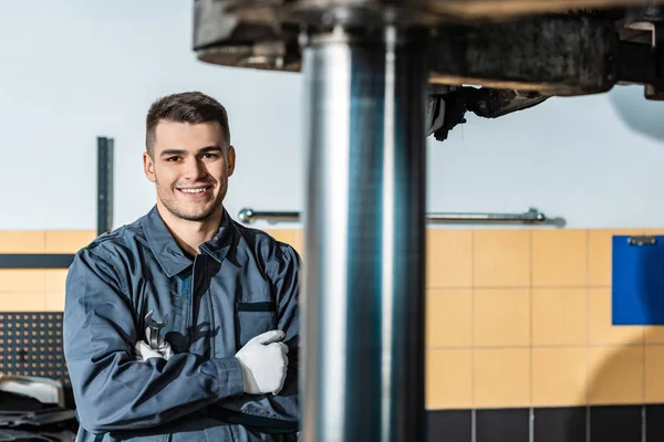 Lächelnder Mechaniker blickt in die Kamera, während er in der Werkstatt neben einem erhöhten Auto steht — Stockfoto