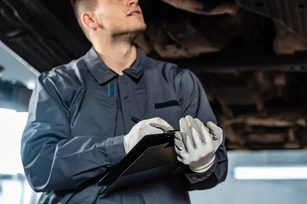 Cropped view of mechanic writing on clipboard while standing under raised car — Stock Photo