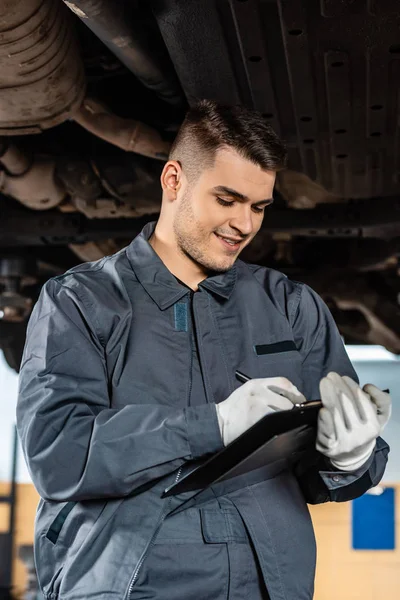Lächelnder Mechaniker schreibt auf Klemmbrett, während er unter erhobenem Auto steht — Stockfoto