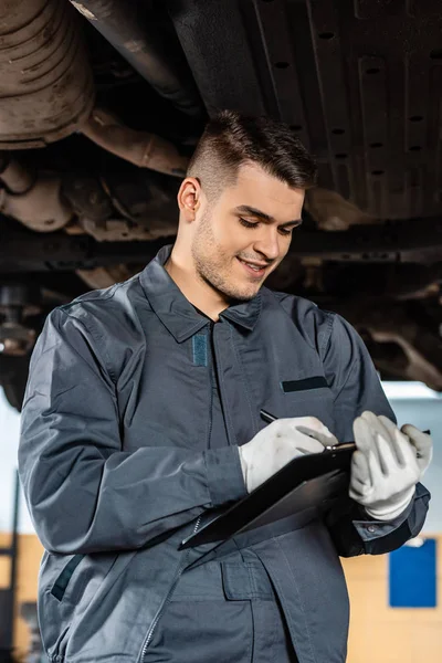 Escritura mecánica sonriente en el portapapeles, mientras que de pie bajo coche elevado - foto de stock