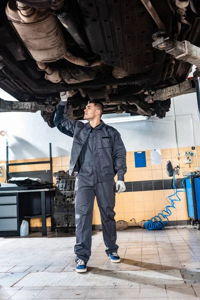 Jeune mécanicien inspectant le fond de la voiture surélevée en atelier — Photo de stock