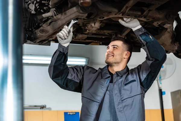 Selective focus of positive mechanic inspecting raised car with wrench — Stock Photo