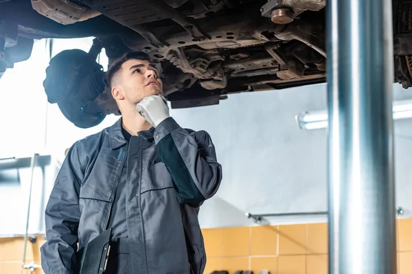 Mécanicien réfléchi inspecter la voiture soulevée sur l'ascenseur de voiture dans l'atelier — Photo de stock