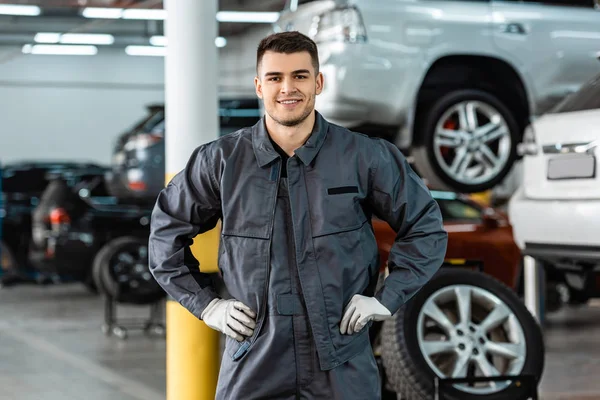 Smiling mechanic looking at camera while standing in workshop with hands on hips — Stock Photo