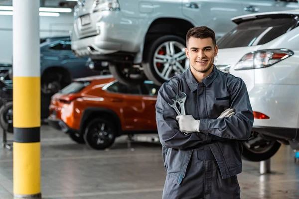 Lächelnder Mechaniker, der mit verschränkten Armen in die Kamera blickt und Schraubenschlüssel hält — Stockfoto