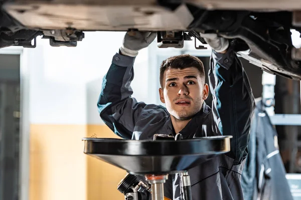 Junger Mechaniker schaut auf den Boden des Autos in der Nähe der Ölförderanlage — Stockfoto