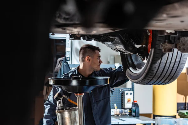 Selective focus of mechanic standing under raised car near waste oil extractor — Stock Photo