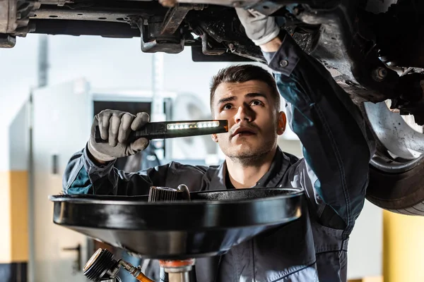 Aufmerksame Mechaniker inspizieren Fahrzeugboden mit Taschenlampe in der Nähe der Altölabsaugung — Stockfoto