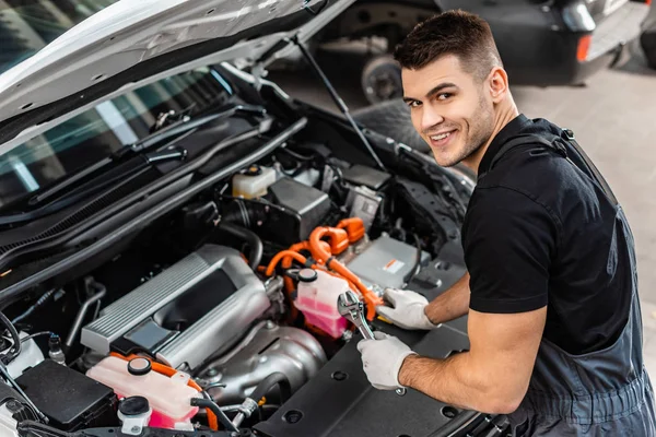 Mecánico guapo inspeccionando el compartimiento del motor del coche y sonriendo a la cámara - foto de stock