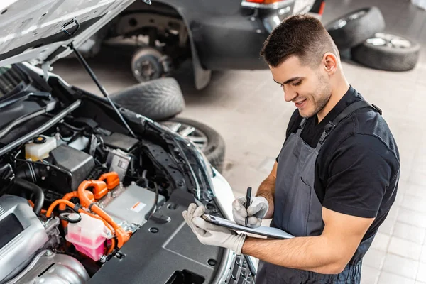 Smiling mechanic writing on clipboard while inspecting car engine compartment — Stock Photo