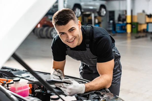 Selektiver Fokus des lächelnden Mechanikers, der auf Klemmbrett schreibt, während er den Motorraum inspiziert — Stockfoto
