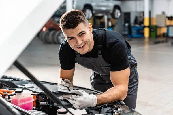 Foyer sélectif de sourire mécanique écriture sur presse-papiers lors de l'inspection du compartiment moteur de la voiture — Photo de stock