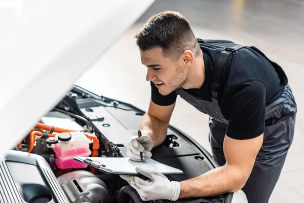 Selective focus of attentive mechanic writing on clipboard while inspecting car engine compartment — Stock Photo