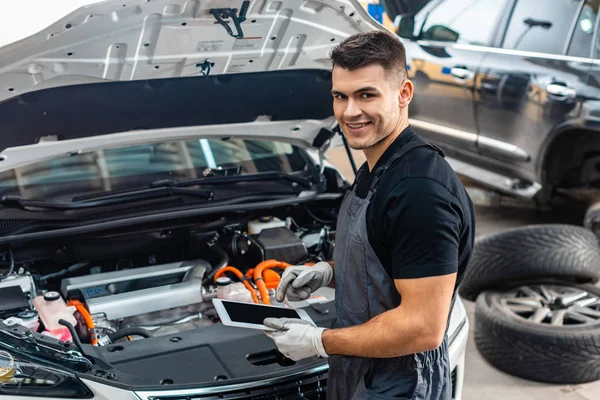 Schöner Mechaniker lächelt in die Kamera, während er ein digitales Tablet in der Nähe des Motorraums benutzt — Stockfoto