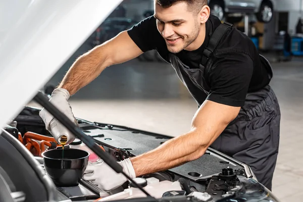 Selective focus of smiling mechanic pouring oil to car engine — Stock Photo