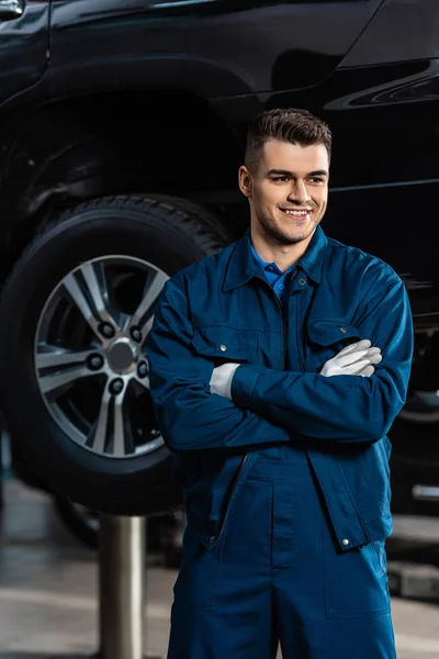 Junger, lächelnder Mechaniker, der mit verschränkten Armen dasteht und wegschaut — Stockfoto