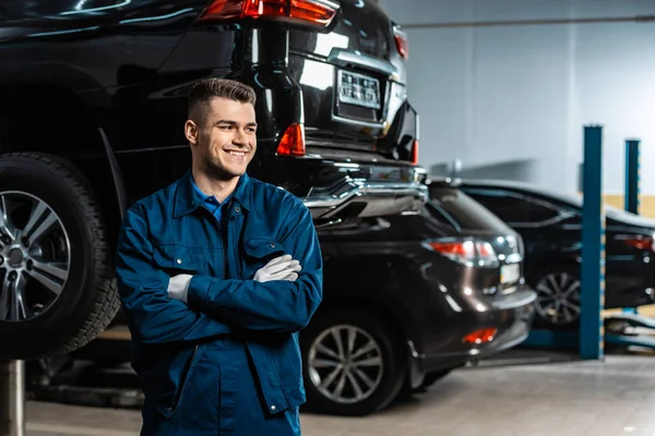 Young, smiling mechanic standing with crossed arms and looking away near modern cars in workshop — Stock Photo