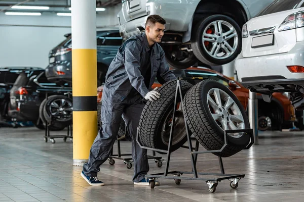 Gutaussehender, lächelnder Mechaniker, der in der Werkstatt neben modernen Autos neue Reifen vom Ständer holt — Stockfoto