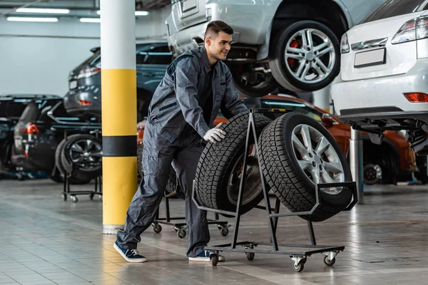 Bonito, sorrindo mecânico tomando novo pneu de stand perto de carros modernos na oficina — Fotografia de Stock