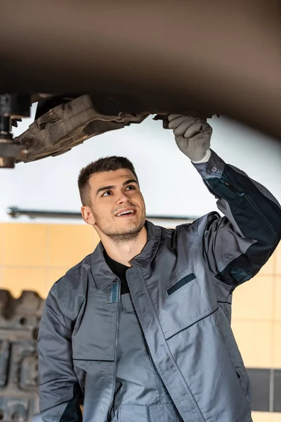 Beau mécanicien inspectant voiture soulevée en atelier — Photo de stock