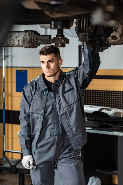 Handsome, serious mechanic standing under raised car and looking away — Stock Photo